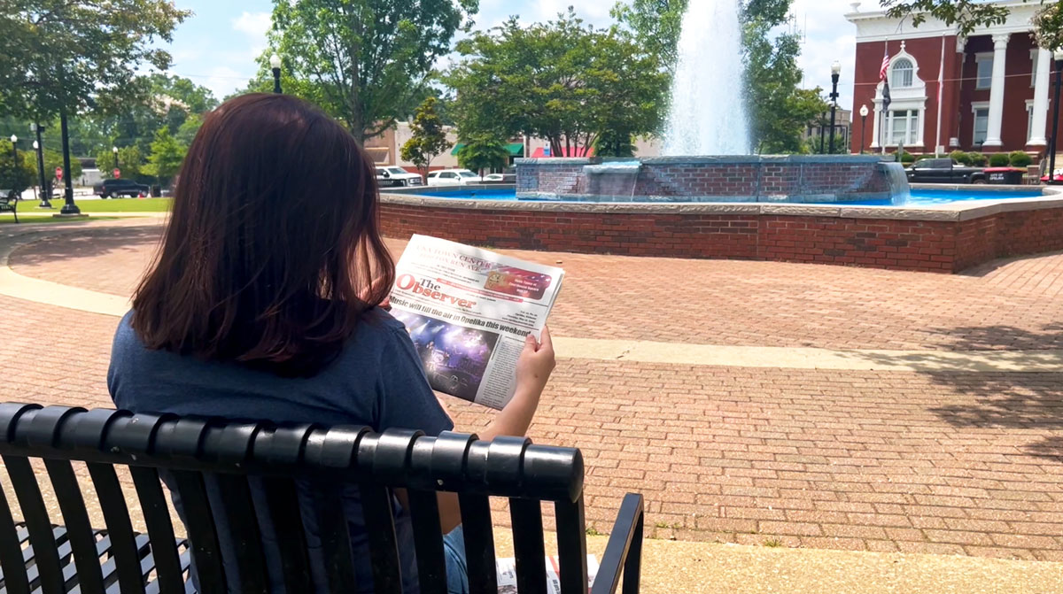 A woman reading the observer on a bench near the Opleika courthouse.